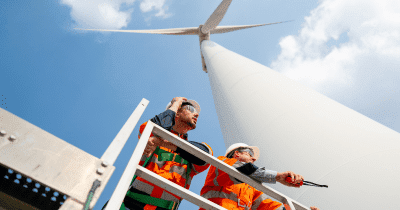 Two Workers Stand On The Base Of A Wind Turbine And Look To Right Side With Blue Sky Aspect Ratio 760 400
